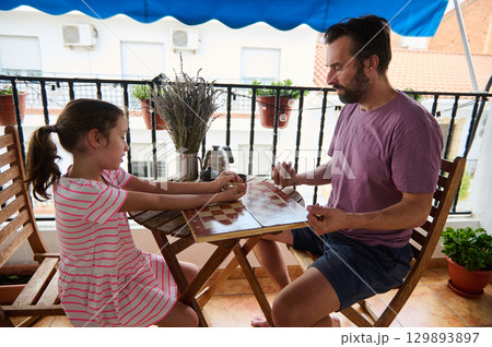 A father and daughter enjoy quality bonding time playing a board game together on a balcony. The secluded space is decorated with potted plants, creating a cozy and welcoming ambiance. A father and daughter enjoy quality bonding time playing a board game together on a balcony. The secluded space is decorated with potted plants, creating a cozy and welcoming ambiance. 129893897