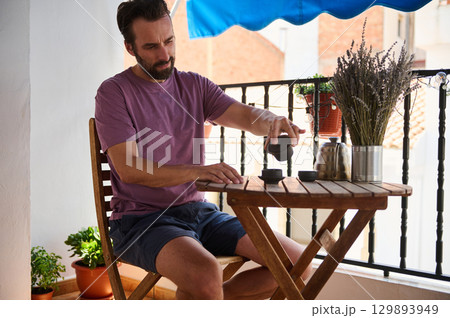 A man sits outdoors on a balcony with wooden furniture, preparing tea. A comfortable and serene setting is complemented by potted plants and a table arrangement, showcasing a calming and peaceful A man sits outdoors on a balcony with wooden furniture, preparing tea. A comfortable and serene setting is complemented by potted plants and a table arrangement, showcasing a calming and peaceful 129893949