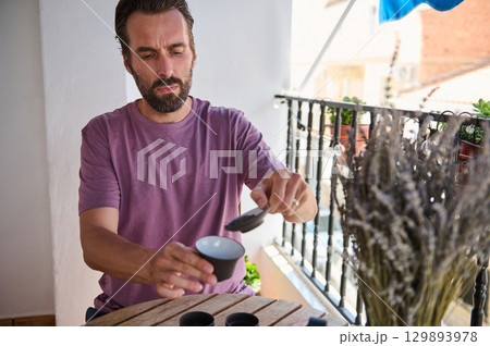 A man performs a tea ceremony on a sunny balcony decorated with plants and lavender. This serene scene highlights personal relaxation, traditional practices, and the enjoyment of a quiet moment. A man performs a tea ceremony on a sunny balcony decorated with plants and lavender. This serene scene highlights personal relaxation, traditional practices, and the enjoyment of a quiet moment. 129893978