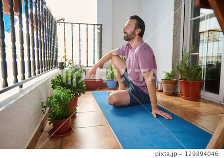 A man enjoys a relaxing yoga session on his balcony, surrounded by potted plants. The bright atmosphere and natural environment promote physical health, mental balance, and tranquility. 129894046