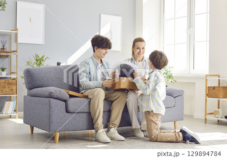 Happy mother with little and teen son unpacking new parcel delivered at home sitting on couch 129894784