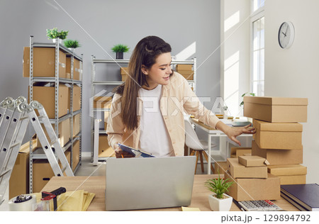 Young confident female employee working in warehouse among cardboard boxes. Young confident female employee working in warehouse among cardboard boxes. 129894792
