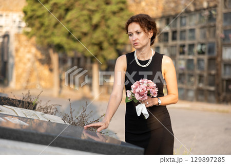 Sad woman in black dress with bouquet of flowers and handkerchief in hands, stands in cemetery Sad woman in black dress with bouquet of flowers and handkerchief in hands, stands in cemetery 129897285