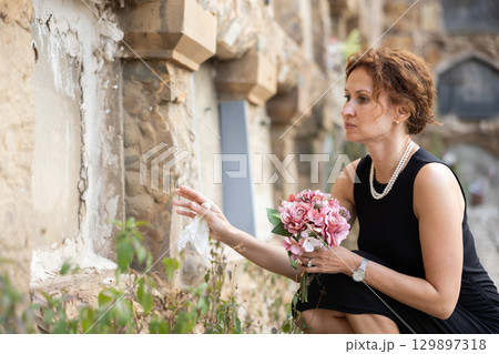 Woman in black crouching in front of columbarium with flowers Woman in black crouching in front of columbarium with flowers 129897318