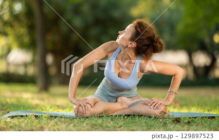 Woman practicing hatha yoga in garden, stretching in an asymmetrical seated asana Parivritta Janu Sirsasana Woman practicing hatha yoga in garden, stretching in an asymmetrical seated asana Parivritta Janu Sirsasana 129897523