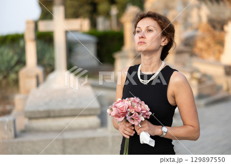 Upset woman in black dress, who lost loved one, brings flowers to grave Upset woman in black dress, who lost loved one, brings flowers to grave 129897550
