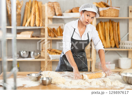 Making dough in bakery - female baker rolling out dough with rolling pin 129897617