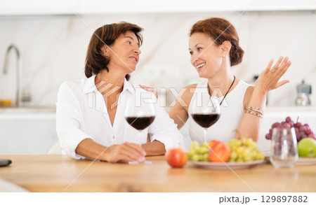 Two girlfriends chatting and drinking red wine in cozy home kitchen 129897882