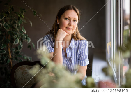 Among indoor plant woman standing near antique armchair and thinking, looking out window Among indoor plant woman standing near antique armchair and thinking, looking out window 129898106