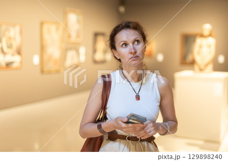 Portrait of woman visitor with smartphone in hands looking exhibition of paintings in museum 129898240