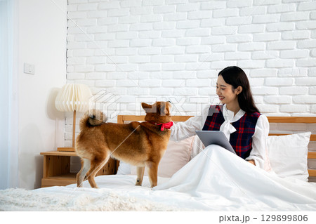 Smiling woman sitting in bed with a tablet, happily petting her dog wearing a red bow in a cozy bedroom Smiling woman sitting in bed with a tablet, happily petting her dog wearing a red bow in a cozy bedroom 129899806