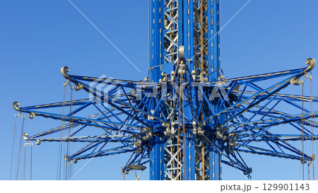 Towering amusement park ride structure with intricate blue metal framework against clear blue sky, showcasing engineering and design excellence Towering amusement park ride structure with intricate blue metal framework against clear blue sky, showcasing engineering and design excellence 129901143
