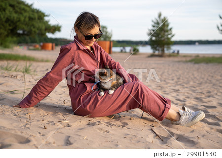 Cat watching woman owner relaxing on sandy beach on seaside walk. Shore stroll with pet feline Cat watching woman owner relaxing on sandy beach on seaside walk. Shore stroll with pet feline 129901530