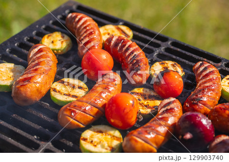 Grilled sausages with vegetables and tomatoes sizzling on barbecue grill outdoors 129903740