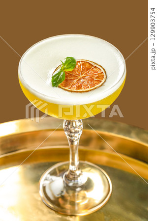 Elegant yellow cocktail in a crystal coupe glass, topped with a frothy foam layer and garnished with a dried citrus slice and fresh basil leaf, served on a golden tray against a brown background 129903754