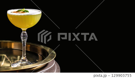 Yellow cocktail with a frothy foam top, garnished with dried citrus and basil leaf, served in an elegant coupe glass on a golden tray against a black background 129903755