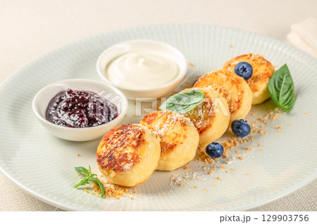 Golden brown syrniki cottage cheese pancakes served on a plate with powdered sugar, blueberries, basil leaves, and bowls of sour cream and berry jam 129903756