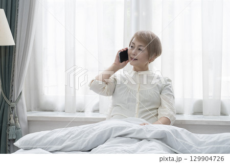 寝室のベッドでスマホを操作するシニア女性 129904726