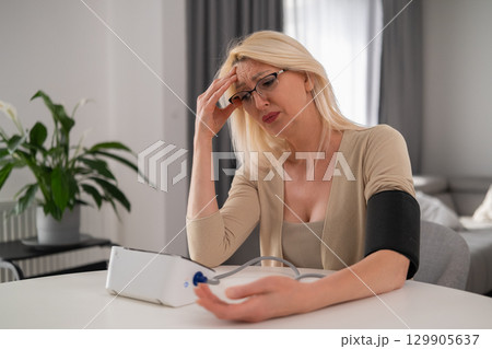 Blonde middle aged woman in beige clothes holding blood pressure monitor, appearing uncomfortable and worried due to headache in modern living room 129905637