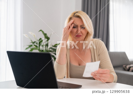 Portrait of blonde middle aged woman sitting in cozy living room, sadly checking bill and struggling with household budget during financial crisis 129905638