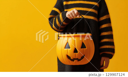 Child black sweater yellow stripe holding pumpkin bucket yellow background Halloween trick or treat festive mood 129907124