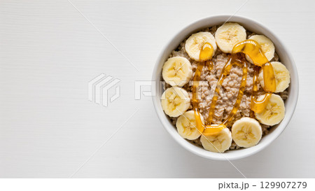 Bowl of oatmeal with banana slices and honey on top, minimalistic breakfast concept on white background 129907279