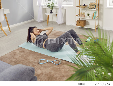Focused sporty woman doing sit ups on yoga mat at home, strengthening body during fitness workout. Focused sporty woman doing sit ups on yoga mat at home, strengthening body during fitness workout. 129907973
