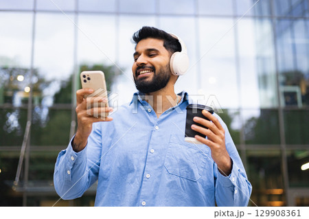 A smiling man listens to music on headphones while looking at his phone and holding a coffee cup. A smiling man listens to music on headphones while looking at his phone and holding a coffee cup. 129908361