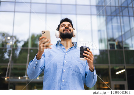 An Indian man in a blue shirt wearing headphones holds a phone and a coffee cup in front of a modern glass building. An Indian man in a blue shirt wearing headphones holds a phone and a coffee cup in front of a modern glass building. 129908362