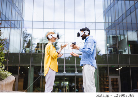 Two people wearing VR headsets stand outdoors in front of a modern building, gesturing as if in a virtual environment. 129908369