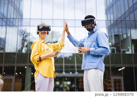 Two people wearing VR headsets high-fiving, standing in front of a modern building, symbolizing technology and teamwork. 129908370