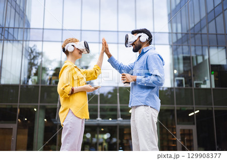 Two people wearing VR headsets give each other a high five outside a modern office building. 129908377