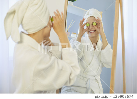 Woman in bathrobe making face mask putting slices of cucumbers on eyes and looking at mirror Woman in bathrobe making face mask putting slices of cucumbers on eyes and looking at mirror 129908674