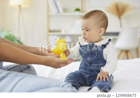 Curious baby in denim overalls reaching for yellow rubber duck toys during playtime with mother. Curious baby in denim overalls reaching for yellow rubber duck toys during playtime with mother. 129908675