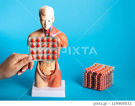 Female hand holding a blister with red pills against the background of a human model's chest, blue background. Cough and pneumonia pills concept, human lung restoration. Copy space for text 129909832