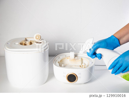 Man in blue gloves spraying descaler, mold and odor remover into humidifier on white background. Humidifier device maintenance concept. Copy space for text 129910016