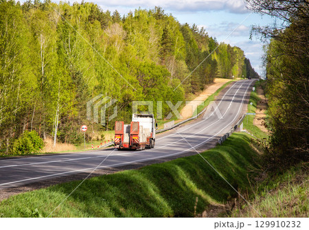 Modern truck with a lowboy semitrailer transports concrete blocks along a country road against the backdrop of a forest in summer. Beautiful nature, industry. Copy space for text 129910232