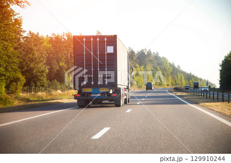 Transportation of a sea metal container on a container ship trailer with a tractor against the background of a forest and a highway in summer. Copy space for text Transportation of a sea metal container on a container ship trailer with a tractor against the background of a forest and a highway in summer. Copy space for text 129910244