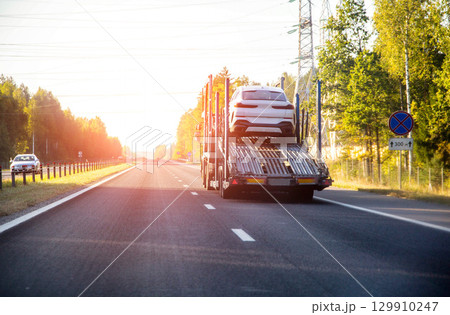 A truck with a semi-trailer for transporting cars on a country road in summer against the backdrop of a sunset. Concept international delivery of cars by car carrier. Copy space for text A truck with a semi-trailer for transporting cars on a country road in summer against the backdrop of a sunset. Concept international delivery of cars by car carrier. Copy space for text 129910247