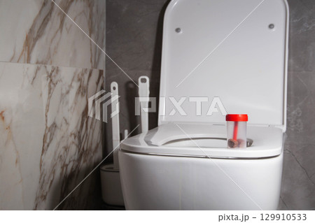Sterile container with stool test on the toilet bowl in the toilet. Concept of stool examination for oviworm and coprogram. Copy space for text, biochemical Sterile container with stool test on the toilet bowl in the toilet. Concept of stool examination for oviworm and coprogram. Copy space for text, biochemical 129910533