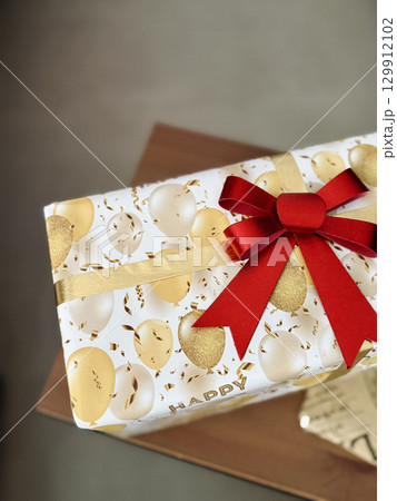 Closeup of festive gift box with gold white balloon print wrapping paper and red bow ribbon, birthday present on wooden table 129912102
