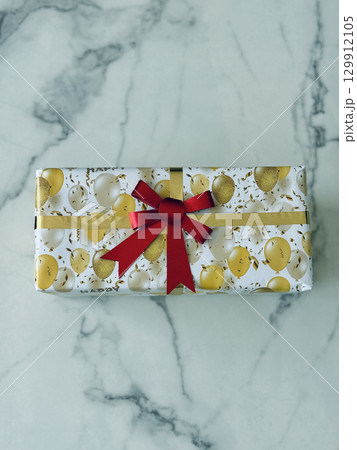 Rectangular gift box wrapped in gold and white balloon print paper with red bow ribbon, festive birthday present on marble surface 129912105