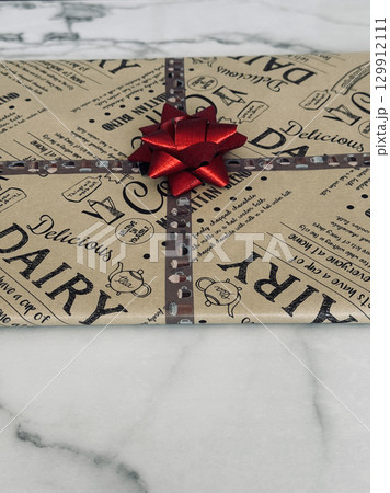 Closeup of gift package with coffee dairy print wrapping paper and red ribbon bow, creative present decor on marble surface 129912111