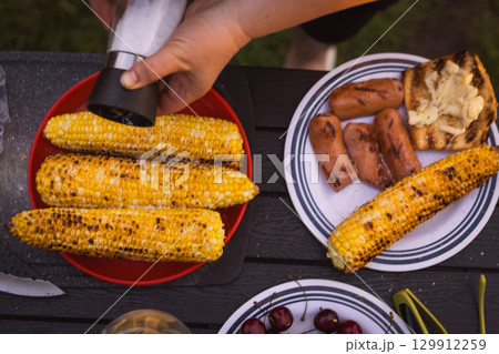 Delicious barbecue spread with grilled corn, sausages, cherries, ready for a summer gathering. A person seasons grilled corn on the cob on a table 129912259