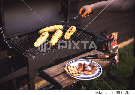 Grilled corn on the cob and sausages are ready for a summer barbecue party. A person grilling corn on the cob and sausages on a barbecue grill outdoors on a wooden table, enjoying a summer meal. 129912260
