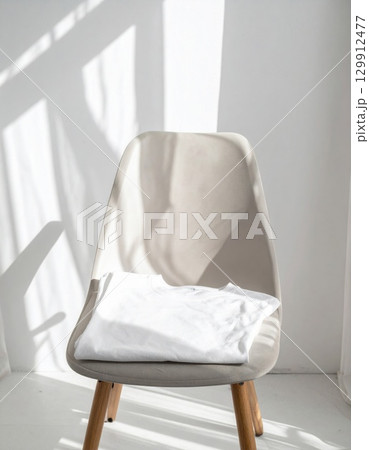 White T Shirt Neatly Folded on Light Beige Chair in a Bright Room with Shadows 129912477