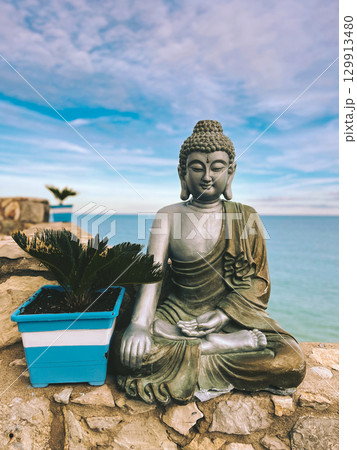 A peaceful Buddha statue meditating by the ocean with a tranquil blue sky as a backdrop. A serene Buddha statue sits in peaceful meditation by the sea, with a small plant adding a touch of nature. 129913480