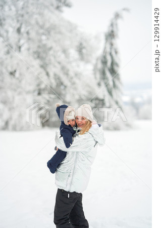 Mother and child in winter clothing enjoying a snowy day outdoors. The mother smiles warmly while holding her bundled-up child, creating a cozy and joyful winter moment Mother and child in winter clothing enjoying a snowy day outdoors. The mother smiles warmly while holding her bundled-up child, creating a cozy and joyful winter moment 129914429
