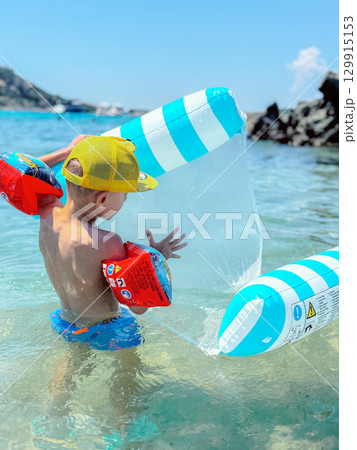 Young child enjoys a sunny day in the ocean, playing with a blue and white inflatable boat. Wearing colorful swim trunks and arm floaties, the child explores and splashes in shallow water. 129915153