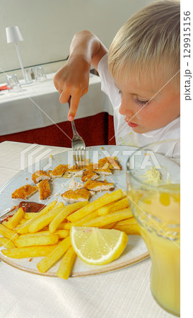 Young child intently eating breaded chicken and French fries at a restaurant. The focus is on the child's enjoyment and concentration while using a fork to interact with the meal. 129915156